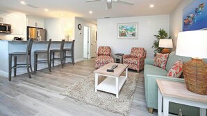 A modern living room with a sofa, chairs, a coffee table, and a dining area with chairs and a kitchen in the background. - Breezy Unit G1 · Sanibel Arms West | 2 Minute Stroll to the Gulf (Sanibel)
