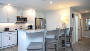 A modern kitchen with white cabinetry, a central island with bar stools, a microwave, and a refrigerator. - Breezy Unit G1 · Sanibel Arms West | 2 Minute Stroll to the Gulf (Sanibel)
