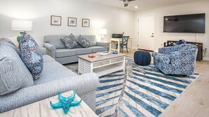 Una sala de estar moderna con un sofá gris, un sillón azul estampado, una mesa de centro y un televisor montado en la pared.