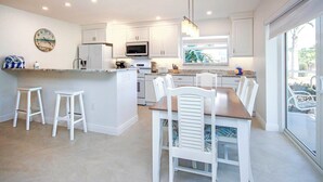 A modern kitchen with white cabinetry, a central island with stools, a dining area with white chairs, and a large window offering a view.