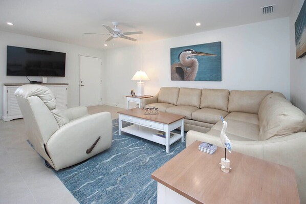 A living room with a beige sofa, a white armchair, a wooden coffee table, and a television on the wall. - Sunny Unit G4 · Sanibel Arms West | Quick 2 Minute Walk to the Beach (Sanibel)