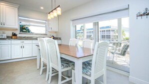 A modern kitchen with white cabinetry, a wooden dining table, and a view of the outdoors through large windows.