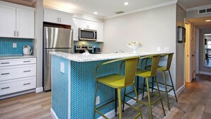 A modern kitchen with white cabinets, a stainless steel refrigerator, a microwave, and a herringbone-patterned backsplash. There are two green bar stools at the island. - Sanibel Vacation Rental | 2-Min Walk to Beach | Sanibel Arms West Condo (Sanibel)