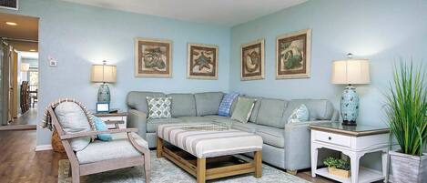 A living room with a grey sofa, a wooden chair, a striped ottoman, and a side table with a lamp. There are framed pictures on the wall and a potted plant on the side table.