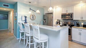 A kitchen with a central island, white bar stools, stainless steel appliances, and a blue accent wall. - Serene Unit G8 Sanibel Arms West Condo | Easy 2 Minute Walk to the Beach (Sanibel)