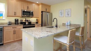 A kitchen with wooden cabinets, stainless steel appliances, a granite countertop, and a small island with two wicker chairs. - Corner Unit with Gulf View (Sanibel)