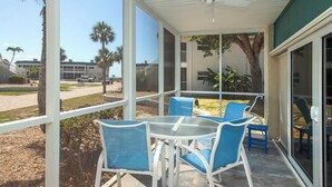 A covered outdoor area with a table and chairs, a ceiling fan, and a view of a parking lot and buildings in the background. - Corner Unit with Gulf View (Sanibel)