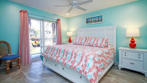 A bedroom with a bed, a chair, a nightstand, a lamp, a ceiling fan, and a window with curtains. - Corner Unit with Gulf View (Sanibel)