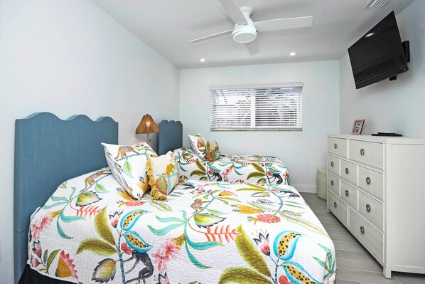 Une chambre à coucher comprenant un lit, une commode, un ventilateur de plafond, un téléviseur et une fenêtre avec des stores.