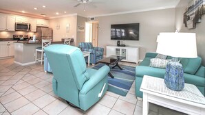A modern living room with a teal sofa, a coffee table, and a TV. There is a kitchen area with a microwave and refrigerator. - Peaceful Condo with Private Beach Access at Sanibel Arms West (Sanibel)