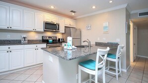 A modern kitchen with white cabinets, granite countertops, a central island with bar stools, stainless steel appliances, and a clock on the wall. - Peaceful Condo with Private Beach Access at Sanibel Arms West (Sanibel)
