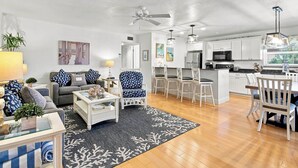 A modern living room with a sofa, coffee table, and a dining area with chairs and a table. There is a kitchen with white cabinets and appliances.