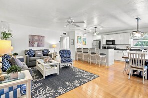A modern living room with a sofa, coffee table, and a dining area with chairs and a table. There is a kitchen with white cabinets and appliances.
