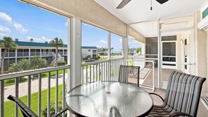 A balcony with a glass table and chairs, a ceiling fan, and a view of a building and palm trees.