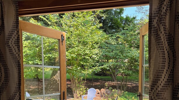 A wooden deck with a glass door leading to a garden area with a white chair and a wooden bench. - Private & Relaxing backyard Hot Tub, Fire pit, nap under the trees in a Hammock (Canadensis)