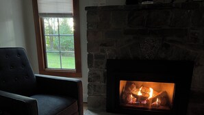 A cozy room with a stone fireplace, a comfortable armchair, and a window with a view of greenery. - Private & Relaxing backyard Hot Tub, Fire pit, nap under the trees in a Hammock (Canadensis)