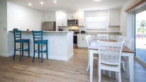 A modern kitchen with white cabinets, a central island with bar stools, a dining table with chairs, and a large window with blinds. - Tranquil Sanibel Arms West Garden Unit J4 | Lush Gardens, Quick Walk to Beach (Sanibel)
