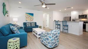 A modern living room with a sofa, a patterned armchair, and a coffee table. There is a kitchen area with a microwave and a refrigerator. - Tranquil Sanibel Arms West Garden Unit J4 | Lush Gardens, Quick Walk to Beach (Sanibel)