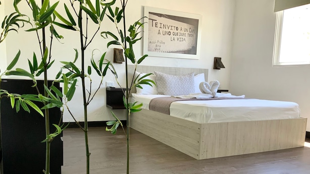 A modern bedroom with a large bed, a bedside table, a wall-mounted lamp, and a framed quote on the wall. - MOKA HOUSE (Santo Domingo)