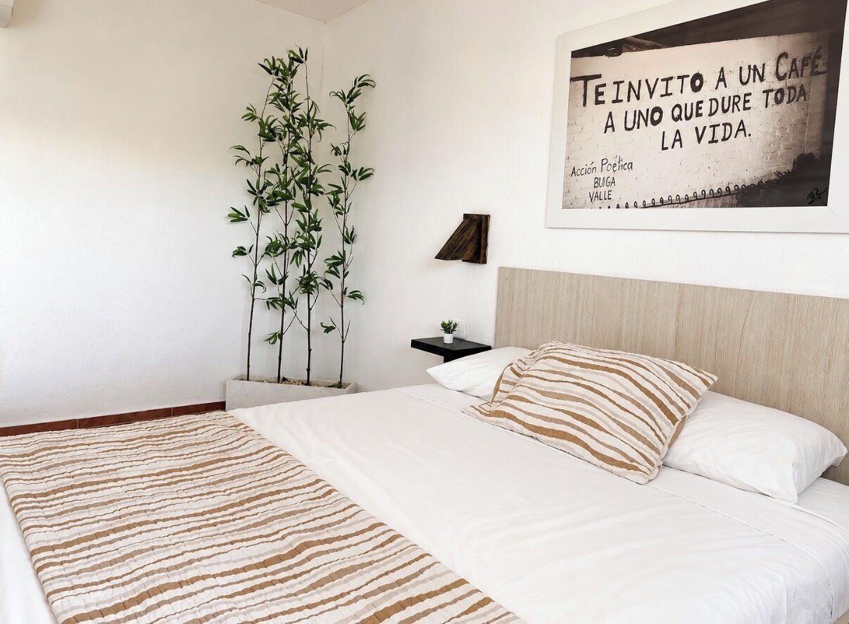 Un dormitorio con cama, una planta colgante, una mesita de noche con lámpara y una cita enmarcada en la pared.