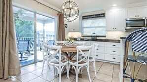 Une cuisine moderne avec des meubles blancs, une table à manger en bois entourée de chaises blanches, un luminaire suspendu et une vue sur l’extérieur grâce à une porte-fenêtre coulissante.