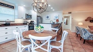Une cuisine moderne avec des meubles blancs, un coin repas doté d’une table ronde et de chaises blanches, et un salon avec des fauteuils à motifs.