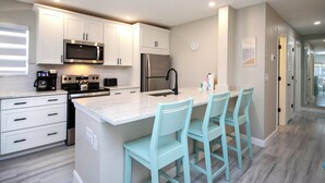 A modern kitchen with white cabinets, stainless steel appliances, a marble countertop, and light blue bar stools.