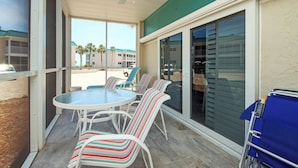 A covered patio area with a round table and striped chairs, a ceiling fan, and a view of a building with palm trees in the background.