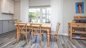 Un coin repas avec une table et des chaises en bois, une vue sur l’extérieur grâce à de grandes fenêtres et un tableau accroché au mur.