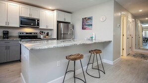 A modern kitchen with white cabinets, stainless steel appliances, a marble countertop, and two wooden stools. - Cozy Unit L3 · Sanibel Arms West Condo | Quick 2 Minute Walk to the Beach (Sanibel)