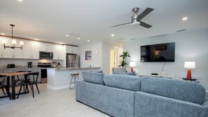 A modern kitchen with white cabinets, a dining area with a wooden table and chairs, a grey sofa, and a ceiling fan. - Cozy Unit L3 · Sanibel Arms West Condo | Quick 2 Minute Walk to the Beach (Sanibel)