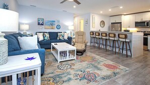 A modern living room with a blue sofa, a white coffee table, and a beige armchair. There is a kitchen area with a bar and a refrigerator. - Tropical Luxury & Poolside Perfection at Sanibel Arms West M2 (Sanibel)