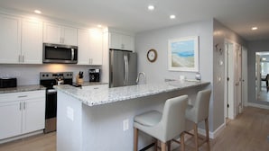 A modern kitchen with white cabinets, stainless steel appliances, a granite countertop, and a central island with bar stools. - Relax in Peaceful Paradise Unit M3 Steps to the Beach (Sanibel)