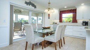 Un coin repas avec une table en verre et quatre chaises, une cuisine avec des meubles blancs et une machine à café, et une porte-fenêtre coulissante donnant sur un patio extérieur.