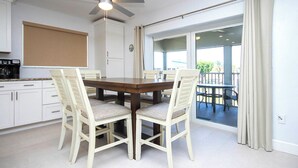 Un coin repas avec une table en bois et des chaises blanches, un ventilateur de plafond et une vue sur un balcon à travers des portes vitrées.