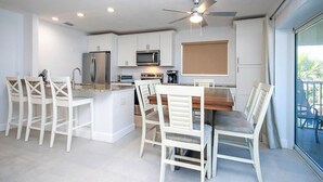 Une cuisine moderne avec des meubles blancs, un îlot central doté de tabourets de bar, un ventilateur de plafond et une grande fenêtre offrant une vue sur l’extérieur.