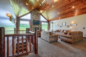 A spacious living room with a stone fireplace, wooden ceiling, and large windows offering a view of the outdoors. - Meadows | Hocking Hills, Pool, Theater, Sleeps 24 (Rockbridge)