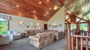 Un vaste salon doté d’un haut plafond en bois, de plusieurs coins salon comprenant des canapés et des fauteuils, et offrant une vue sur la verdure environnante.