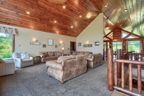 A spacious living room with a high wooden ceiling, multiple seating areas including sofas and armchairs, and a view of greenery outside.