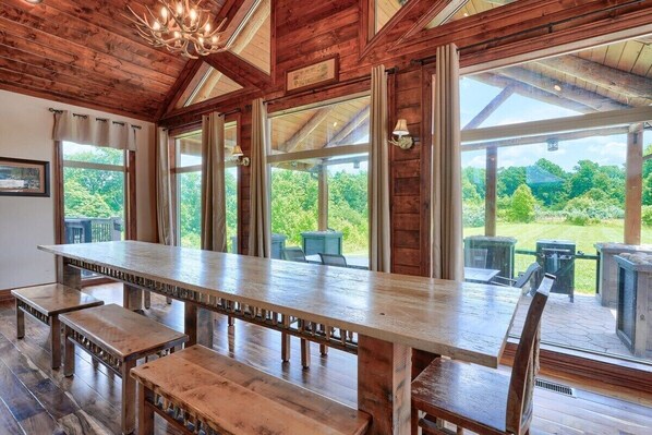 A wooden dining area with a long table and benches, large windows with curtains, and outdoor seating visible through the windows. - Meadows | Hocking Hills, Pool, Theater, Sleeps 24 (Rockbridge)