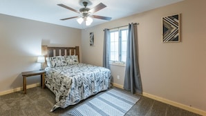 A bedroom with a bed, a nightstand, a ceiling fan, a window with curtains, and a wall art piece. - Wolf Den | 6BR, Indoor Pool, Arcade, Pond, Pets (Logan)