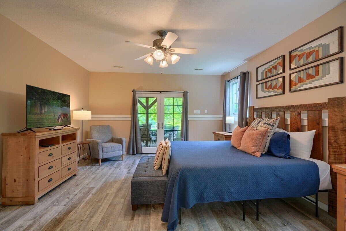 Ein Schlafzimmer mit einem Bett, einem Sessel, einer Holzkommode, einem Deckenventilator und einem Fenster mit Blick auf Grünflächen.