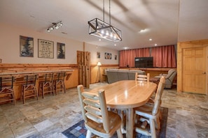 A cozy dining area with a wooden table and chairs, a bar with stools, and a television in the background.
