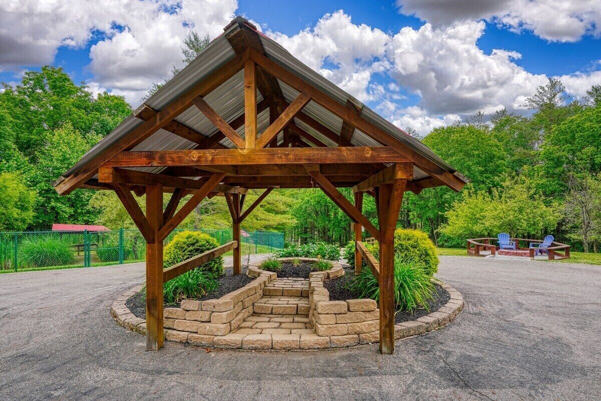 A wooden gazebo with a metal roof, surrounded by a landscaped garden and a paved area with seating.