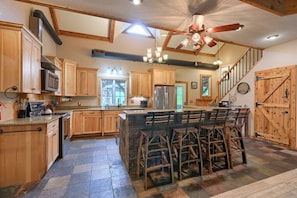 A spacious kitchen with wooden cabinets, a central island with bar stools, a stainless steel refrigerator, and a ceiling fan.