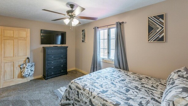 Une chambre à coucher comprenant un lit, une commode, un ventilateur de plafond, une fenêtre avec des rideaux et un téléviseur fixé au mur.