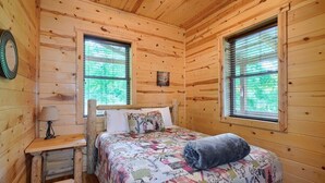 Un dormitorio de cabaña de madera con una cama, dos ventanas, una mesita de noche y una lámpara.