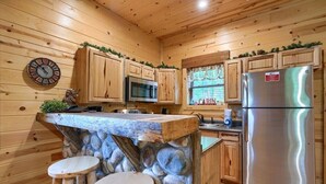 Cocina tipo cabaña con paredes de madera, una isla de piedra, dos taburetes, un refrigerador de acero inoxidable y un reloj.
