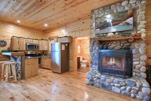 A wooden cabin interior with a stone fireplace, kitchen area, and a stone wall with a TV.