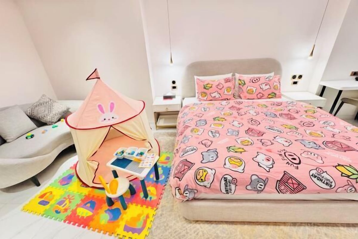 Une chambre avec un lit, un ciel de lit rose, un ordinateur portable et un tapis de jeu coloré.